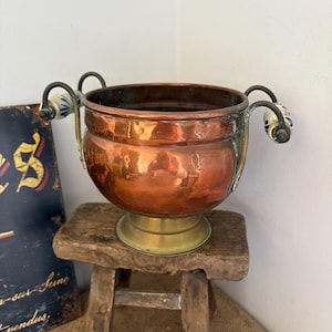 May include: A round copper bowl with a polished finish, featuring a wide opening and a brass pedestal base. The bowl has two curved metal handles on opposite sides, each accented with a white ceramic grip decorated with a blue floral pattern.