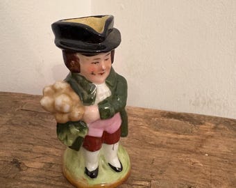 May include: A ceramic figurine of a person wearing a black tricorne hat, a dark green coat, pink breeches, and white stockings. The figure holds a cluster of white flowers in their arms and stands on a small, circular, light green base.