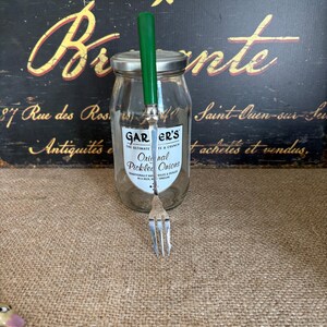 May include: A glass jar of "GAR ER'S Original Pickled Onions" with a silver lid and a green-handled fork. The jar is set against a vintage sign with French text. The fork is approximately 6 inches long.