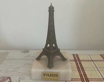 Könnte beinhalten: Eine kleine Metall-Replik des Eiffelturms auf einem weißen Marmorsockel mit den Worten "PARIS" und "www.creativeparis.com" auf einem goldenen Etikett.