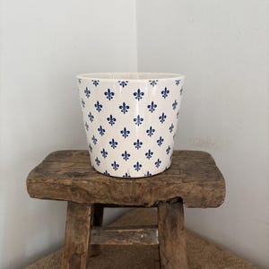 Może przedstawiać: Biała ceramiczna donica z powtarzającym się wzorem niebieskich symboli fleur-de-lis. Donica jest cylindryczna i stoi na małym, rustykalnym drewnianym stołku. Stołek ma kwadratowy blat i cztery nogi.