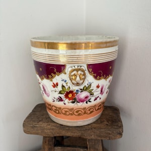 May include: A porcelain cachepot featuring a white base with a gold rim, a deep magenta band, and a central relief lion head motif. The body is decorated with hand-painted floral arrangements in pink, red, and green, finished with a gold scroll border.