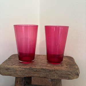 May include: Two tall, clear drinking glasses in a vibrant magenta colour. The glasses are cylindrical with a slightly flared top. They are set on a rustic, weathered wooden stool against a plain white wall. The glasses are empty.
