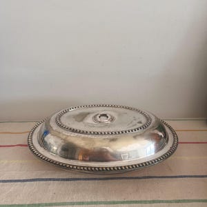 Puede incluir: Una fuente ovalada de servir de plata con tapa. La fuente tiene un borde con cuentas y un pequeño pomo en la tapa.