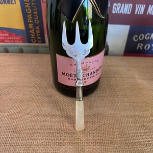 以下が含まれることがあります： 3 本の歯を持つフォークのデザインが施された、銀と真珠貝のシャンパンボトルストッパー。ストッパーは、モエ・エ・シャンドン ロゼ アンペリアル シャンパンのボトルの隣にある麻の表面に置かれています。