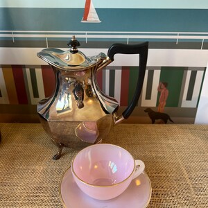 Może przedstawiać: Srebrny czajnik z czarnym uchwytem i trzema nóżkami, obok różowego kubka do herbaty i spodeczka z złotą obwódką.