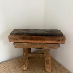 Könnte beinhalten: Ein rechteckiger Schleifstein in einem verwitterten Holzrahmen, der auf einem kleinen, rustikalen Holzhocker mit vier dicken, stabilen Beinen ruht, wobei eine horizontale Strebe die Beine unter der Sitzfläche verbindet.