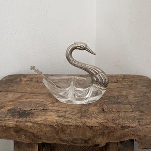 May include: A clear glass swan-shaped serving dish with a silver-colored metal swan head and neck. The dish has three compartments and sits on a rustic wooden surface. A small spoon rests on the side of the dish.