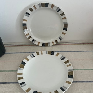 May include: Two white ceramic plates with a black, brown and silver geometric pattern around the rim.