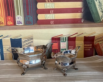 May include: Two silver-plated serving dishes with ornate handles and feet are displayed on a striped surface. The background features a collection of vintage books with colorful spines, including French and English titles.