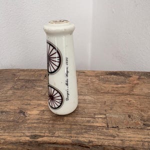 Peut inclure: Une poignée cylindrique en céramique blanche ornée d'une illustration graphique d'un véhicule ancien à roues à rayons. Le texte "Duryea's Motor Wagon, 1895" est imprimé en écriture noire sur le côté de la surface lisse et brillante.