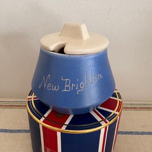 Puede incluir: Un azucarero de cerámica azul con una tapa blanca y las palabras "New Brighton" escritas en letras doradas. El azucarero está sobre una caja azul y roja con un borde dorado y una corona.