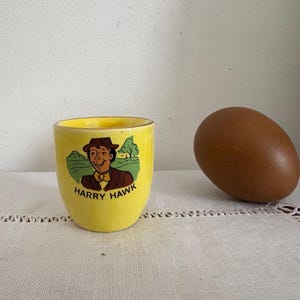 May include: Yellow ceramic egg cup with a cartoon illustration of a man wearing a hat and a bow tie. The text "HARRY HAWK" is printed on the cup.