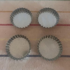 May include: Four small, round, fluted tart pans made of silver-coloured metal. The pans are arranged in a square on a striped, neutral-toned fabric. The edges of the pans have a scalloped design.