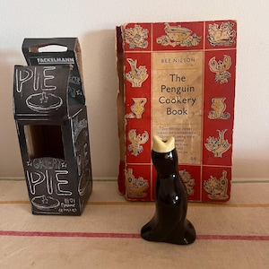 May include: A vintage cookbook titled "The Penguin Cookery Book" by Bee Nilson. The book cover features a red and white design with illustrations of various foods. A ceramic penguin gravy boat with a yellow crown is in the foreground.