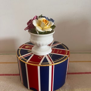 May include: A white ceramic vase with a floral arrangement of yellow, purple, and blue roses sits atop a round box. The box is decorated with a Union Jack design in red, white, and blue, with gold trim.