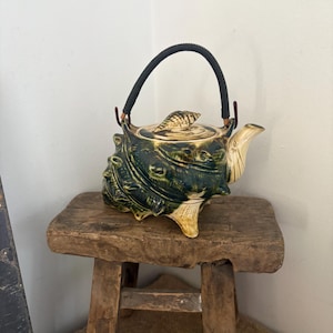 May include: A ceramic teapot with a seashell design, in dark green and beige. It has a black handle and sits on a small, rustic wooden stool. The teapot's design includes textured details. The teapot is a decorative item.