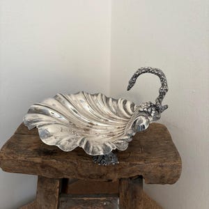 May include: A silver-coloured, shell-shaped dish with a decorative handle. The dish has a ruffled edge and sits on a rustic wooden stool. The handle is ornate and textured, adding a touch of elegance. The dish is likely for serving or display.