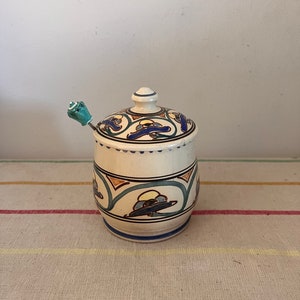 Può includere: Un barattolo in ceramica con coperchio e cucchiaio. Il barattolo è bianco sporco con motivi decorativi blu, gialli e marroni. Il cucchiaio ha un manico turchese. Il barattolo è su un tessuto a righe.