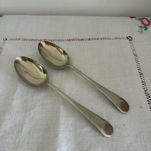 Könnte beinhalten: Zwei silberne Löffel mit ovalen Laffen und langen, schlanken Griffen. Die Löffel liegen auf einer weißen, strukturierten Oberfläche. Die Löffel dienen wahrscheinlich zum Servieren oder Essen.