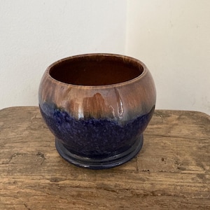 May include: A round ceramic bowl with a dark brown interior and a glossy exterior. The bowl's exterior transitions from a deep blue base to a brown and purple upper section. The bowl sits on a small, circular base.