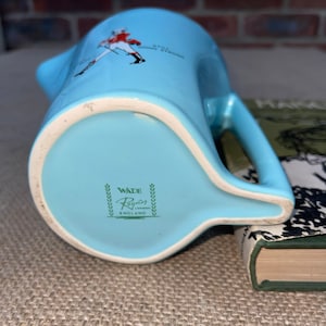 May include: A light blue ceramic jug with a handle. The jug has a green and white logo that reads "Wade Regon London England" and a red and white illustration of a man in a military uniform with the text "Still Going Strong".
