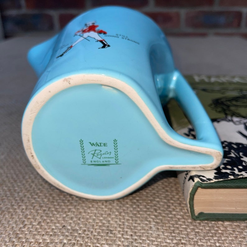 May include: A light blue ceramic jug with a handle. The jug has a green and white logo that reads "Wade Regon London England" and a red and white illustration of a man in a military uniform with the text "Still Going Strong".