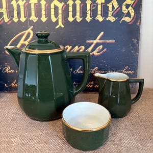 May include: A dark green ceramic tea set with gold edging, comprising a teapot, creamer jug, and sugar bowl. The set is displayed on a textured surface, with a vintage sign in the background.