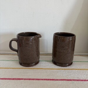Puede incluir: Dos tazas de cerámica marrón. Una tiene asa y boquilla, la otra es una taza cilíndrica. Ambas tienen una textura moteada y un esmalte marrón oscuro. Las tazas están sobre una tela a rayas.