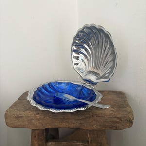 May include: A vintage, blue glass butter dish in the shape of a seashell, with a silver-colored metal lid and trim. A small, silver-colored butter knife rests inside the dish. The dish is open, revealing the blue glass interior.
