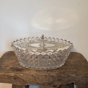 Pode incluir: Uma travessa oval de vidro transparente com tampa. A travessa tem um padrão de corte de diamante nas laterais e uma borda decorativa. A tampa apresenta uma pega prateada e um design estampado. A travessa está colocada numa superfície de madeira rústica.