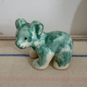 May include: A ceramic bear figurine in shades of green and white. The bear is in a standing position with its head turned slightly to the left. The figurine has small black eyes and rounded ears. The bear is on a striped fabric.