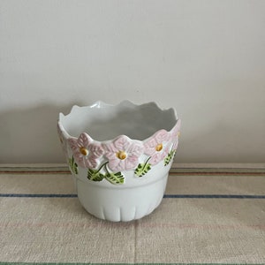以下が含まれることがあります： ピンクと緑の花柄が施された、白いセラミック製のプランター。縁はギザギザになっている。