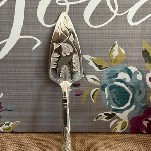 Puede incluir: Una espátula plateada para tartas con un diseño floral ornamentado en la hoja y el mango. La espátula tiene una hoja triangular puntiaguda y un mango detallado. El fondo presenta un patrón floral gris y la palabra "Love" en blanco.