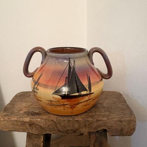 Könnte beinhalten: Eine Keramikvase mit zwei Henkeln, die eine Sonnenuntergangsszene mit Segelbooten zeigt. Die Vase hat eine runde Form mit braunem Innenraum und einem Farbverlauf aus Orange, Gelb und Lila. Die Segelboote sind in Schwarz dargestellt.