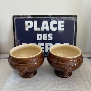 Puede incluir: Dos cuencos de sopa de cerámica marrón con interior beige, colocados sobre una superficie a rayas blancas y azules. Los cuencos tienen asas decorativas y se asientan sobre una base de pedestal. Un letrero azul con el texto blanco "PLACE DES" está en el fondo.