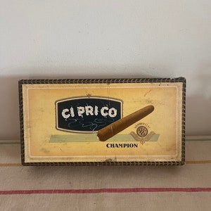 Puede incluir: Una caja vintage de cigarros "Ciprico" con un diseño marrón y dorado. La caja presenta un cigarro con una banda blanca que dice "Vanderlust" y la palabra "Champion" impresa en la parte inferior.