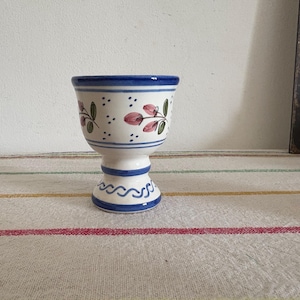 May include: A white ceramic egg cup with a blue rim and base, decorated with pink floral and green leaf designs. The egg cup has a pedestal base and is approximately 4 inches tall.