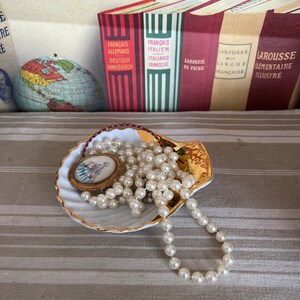 May include: A white seashell-shaped dish with a gold rim, containing a pearl necklace and a small, round, gold-framed picture with a floral design.