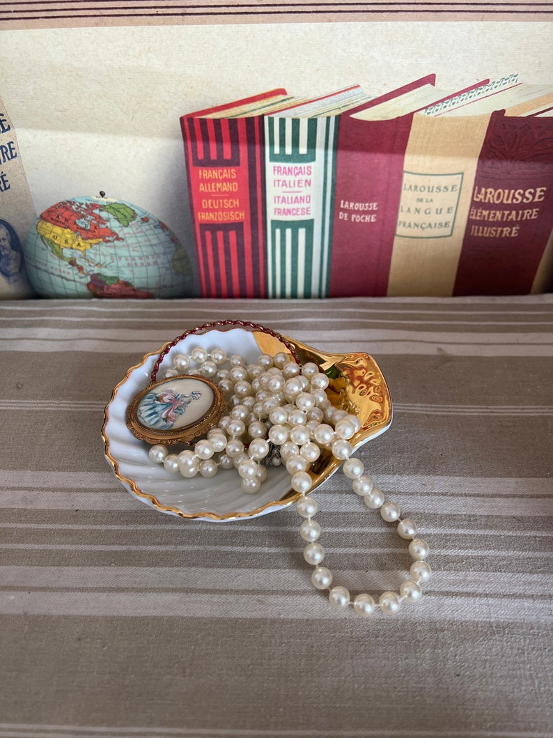 May include: A white seashell-shaped dish with a gold rim, containing a pearl necklace and a small, round, gold-framed picture with a floral design.