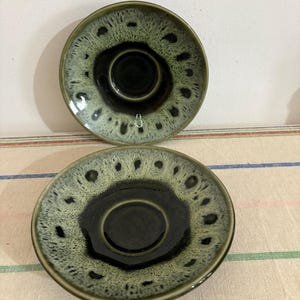 May include: Two green and black ceramic saucers with a speckled glaze. The saucers have a raised rim and a center depression.