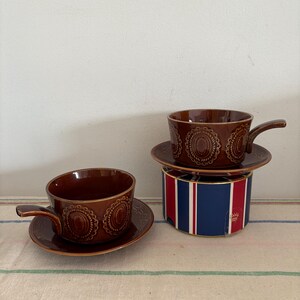 May include: Two brown ceramic cups with matching saucers. The cups have a raised design and a handle. The cups are sitting on a striped surface.