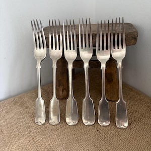 May include: A set of six silver-toned metal forks arranged in a row. Each fork features four tines, a slender neck, and a wide, flat handle with a rounded, heart-shaped base. The metal has a smooth, polished finish with subtle signs of wear.