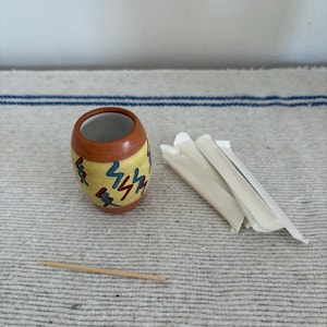 May include: A small, ceramic toothpick holder with a yellow base, brown top, and colorful abstract design. A wooden toothpick and a small stack of wrapped toothpicks are next to the holder.