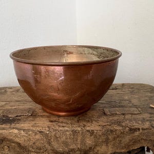 May include: A round copper bowl with a polished exterior and a weathered, tarnished interior surface. The bowl features a smooth, curved silhouette with a slightly flared rim, showcasing the metallic reddish-brown tones of the copper material.