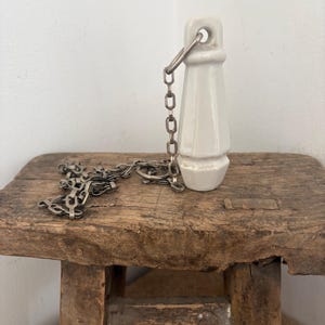 May include: A white ceramic toilet pull with a metal chain, resting on a rustic wooden stool. The pull is cone-shaped with a loop at the top for the chain. The chain is silver-colored and lies across the stool's surface.