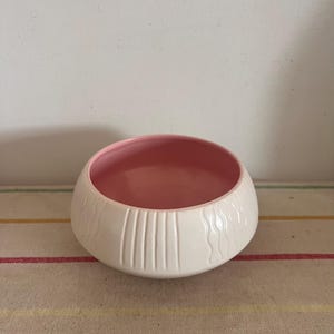 Peut inclure: Un bol en céramique blanc avec un intérieur rose. L'extérieur présente des motifs texturés verticaux et ondulés. Le bol est posé sur une surface en tissu rayé, mettant en valeur son design et son contraste de couleurs. Ce bol décoratif convient à la décoration intérieure.