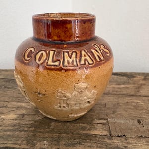 Könnte beinhalten: Antiker Colman's Senftopf. Das Steinzeuggefäß hat einen braun glasierten Rand und den Schriftzug "COLMAN'S" in erhabenen Buchstaben. Auf der Seite befindet sich ein Reliefbild. Der Topf steht auf einer verwitterten Holzoberfläche.