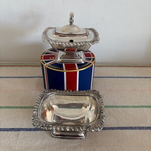 Puede incluir: Dos fuentes plateadas. Una es una fuente adornada con tapa, la otra una fuente rectangular a juego. Se encuentran sobre un recipiente con un diseño de la Union Jack. Las fuentes tienen bordes decorativos.