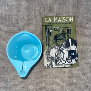 May include: A vintage book titled "La Maison: The History of Prunier's" with a green cover and black illustrations. The book features a woman in a black and white dress and a man in a top hat. The book is next to a light blue ceramic jug with a handle.